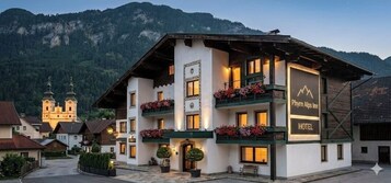 Um hotel com fachada branca, varandas adornadas com caixas de flores e montanhas ao fundo.