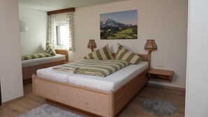 A bedroom with a wooden bed, a window with curtains, a painting of a mountain landscape, and a small bedside table with a lamp.