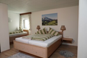 A bedroom with a wooden bed, a window with curtains, a painting of a mountain landscape, and a small bedside table with a lamp.