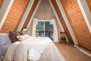 Ein Schlafzimmer mit schrägen Wänden, einem Bett mit vielen Kissen, einem Sessel und einem Fenster mit Vorhängen.