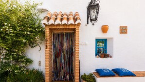 Edifici blanc amb teulada a esquerdes, entrada, porta blava i finestra petita amb plantes en testos.