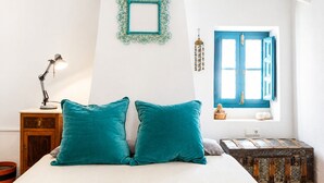 Un llit amb almoinades color turquesa, un escriptori amb una làmpada, un quadre decoratiu penjat a la paret, una finestra amb persianes blaves i un moble de prestatgeries.