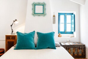 Ein Bett mit türkisblauen Kissen, ein Schreibtisch mit Lampe, ein dekorativer Wandrahmen, ein Fenster mit blauen Rollläden und eine Kommode.