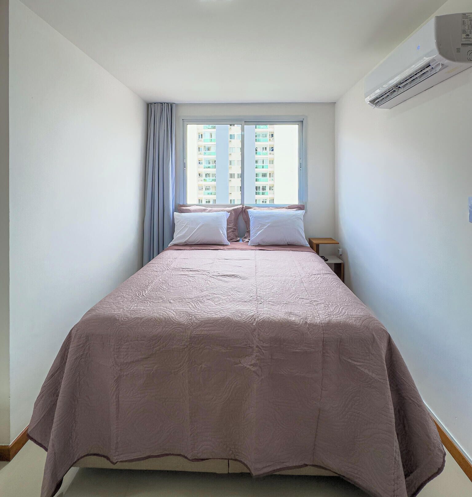 A bedroom with a bed, two pillows, a nightstand, a window with curtains, and an air conditioning unit.