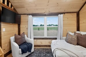 Ein Cabin-Zimmer mit Holzvertäfelung, einer Couch, einem Sessel, einem an der Wand montierten Fernseher und einem Fenster mit Vorhängen.