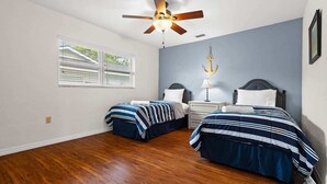 Une chambre avec deux lits, un ventilateur de plafond, une table de chevet avec une lampe, une fenêtre avec des stores et une décoration murale en forme d’ancre.