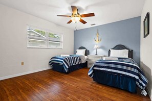 Une chambre avec deux lits, un ventilateur de plafond, une table de chevet avec une lampe, une fenêtre avec des stores et une décoration murale en forme d’ancre.