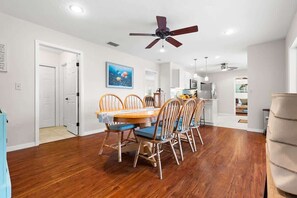 Un coin repas avec une table et des chaises en bois, un ventilateur de plafond et une cuisine en arrière-plan.