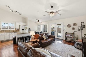 A spacious living area with a brown leather sofa, a dining table with chairs, a kitchen with white cabinets, and a ceiling fan.