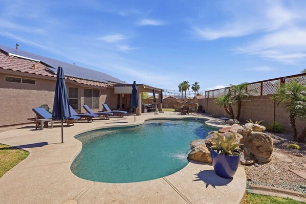 A swimming pool with lounge chairs, a covered patio, and a fenced area with palm trees.