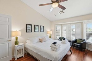A bedroom with a bed, bedside lamps, a ceiling fan, a window with blinds, and a chair.