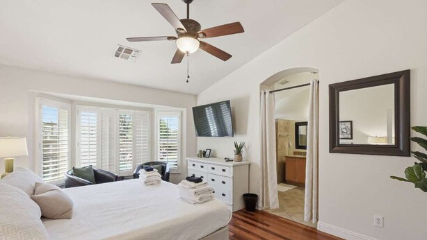Un dormitorio con cama, ventilador de techo, televisión, cómoda y un baño visible a través de un arco.