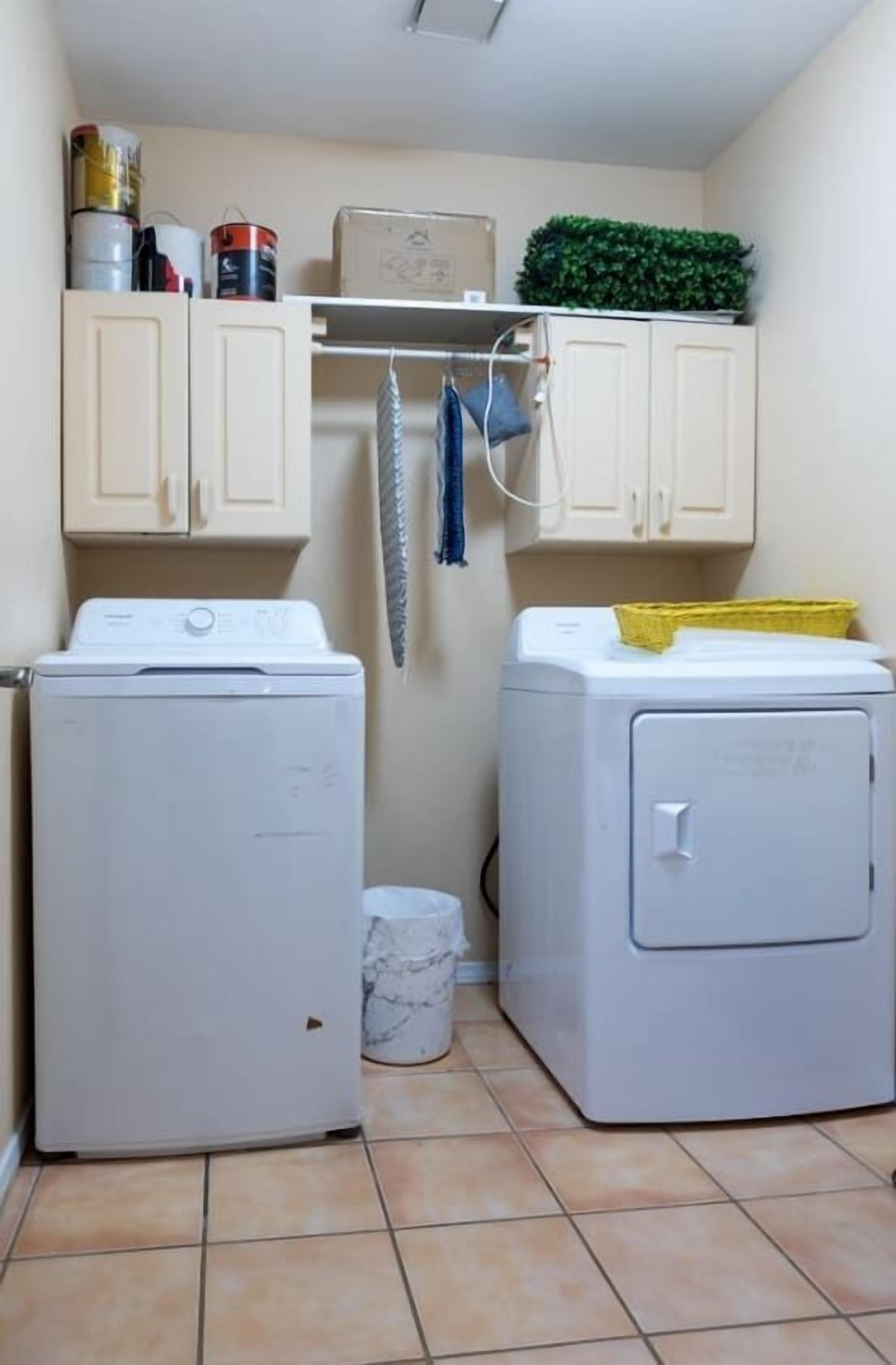 A laundry room with two white washing machines, a marble trash can, and cabinets above holding various items.