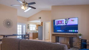Ein Wohnzimmer mit Flachbildfernseher an der Wand, Deckenventilator und Essbereich im Hintergrund.
