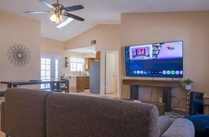 Ein Wohnzimmer mit Flachbildfernseher an der Wand, Deckenventilator und Essbereich im Hintergrund.