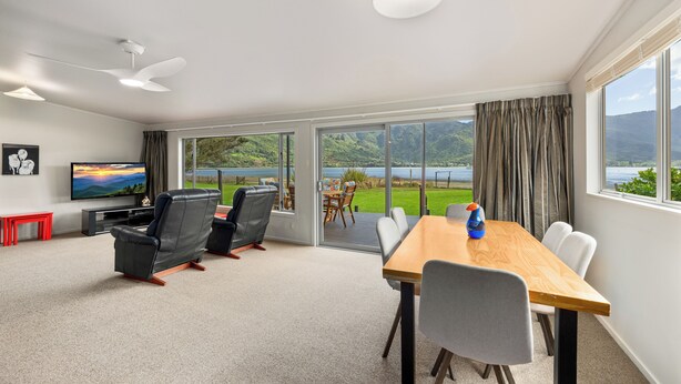 A modern living room with a flat-screen TV, leather chairs, a wooden dining table, and a large sliding glass door leading to a balcony with a scenic view.
