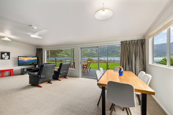 A modern living room with a flat-screen TV, leather chairs, a wooden dining table, and a large sliding glass door leading to a balcony with a scenic view.