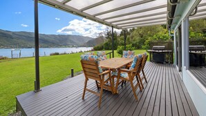 A wooden deck with a table and chairs, overlooking a lake and mountains.