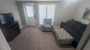 A living room with a television on a stand, a patterned ottoman, a dark sofa, and a door with a window.