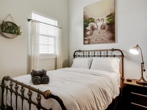 Una cama bien hecha con cabecera, lámpara de noche, planta en la pared y ventana con persianas.