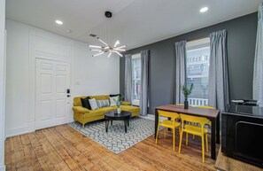 A room with a wooden floor, a yellow sofa, a small table, a TV, and a window with curtains. - Cheerful & Central 1-bd/1-ba w Backyard (Houston)