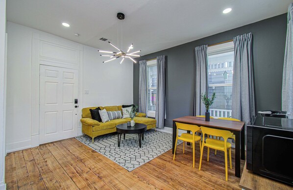 A room with a wooden floor, a yellow sofa, a small table, a TV, and a window with curtains. - Cheerful & Central 1-bd/1-ba w Backyard (Houston)