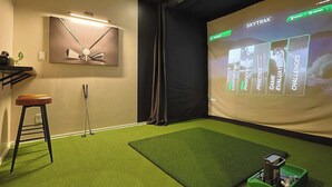A room with a projector screen displaying a game interface, a green artificial turf floor, a stool, and a wall-mounted shelf. - 5-bd/2.5-ba With Pool, Hot Tub, Cinema & Golf Sim (Royse City)