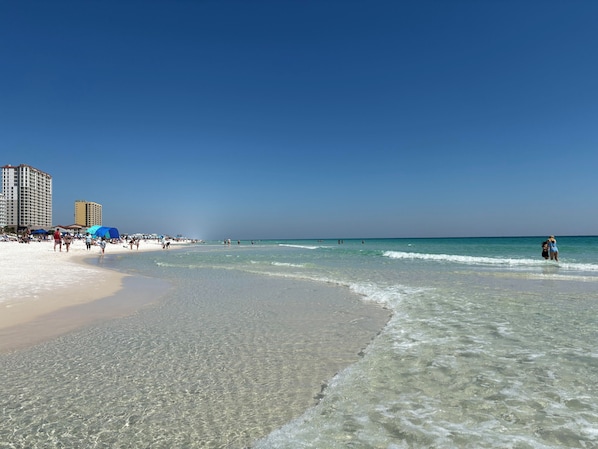 A beach with clear water, umbrellas, and buildings in the background. - Sun, Safari, Shopping- 3 BR Escape, Drive to Beach (Gulf Breeze)