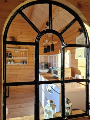 Ein rustikaler Holzinnenraum mit einem großen Bogenfenster, Holzbalken an der Decke und einem durch das Fenster erkennbaren Küchenbereich.