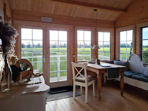 Ein Holzblockhaus-Interieur mit Esstisch, Stühlen, einer Couch und großen Fenstern mit Blick auf eine grüne Landschaft.