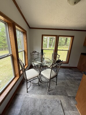 A dining area with a glass table and metal chairs, large windows with a view of trees, and a ceiling with a light fixture.
