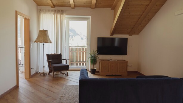 Een woonkamer met een houten plafond, een televisie, een bank, een stoel, een lamp en een balkon met uitzicht op bergen.