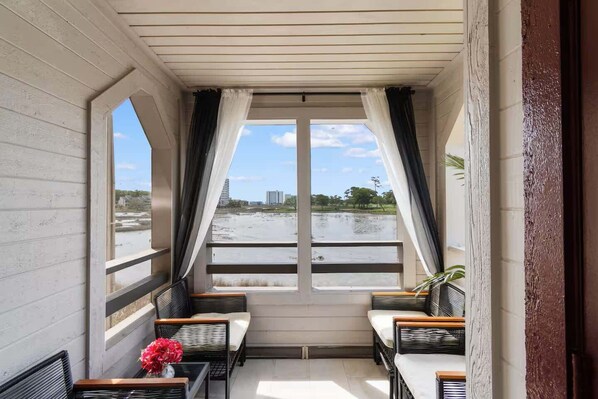A balcony with wicker furniture, a potted plant, and a view of a body of water and buildings. - Luxury Condo, Waterviews, Pools, 12PM Check In (Myrtle Beach)