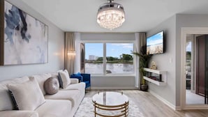 Ein modernes Wohnzimmer mit einem weißen Sofa, einem runden Couchtisch, einer Kandelaberleuchte, einem Fernseher an der Wand und Blick auf einen Strand durch das Fenster.
