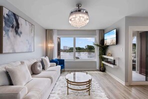 A modern living room with a white sofa, a round coffee table, a chandelier, a TV on the wall, and a view of a beach through the window. - Luxury Condo, Waterviews, Pools, 12PM Check In (Myrtle Beach)
