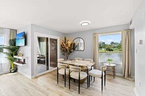 A modern room with a round dining table, chairs, a TV, and a view of a lake. - Waterfront Condo: Marsh View, Pools, 12pm Check-In (Myrtle Beach)