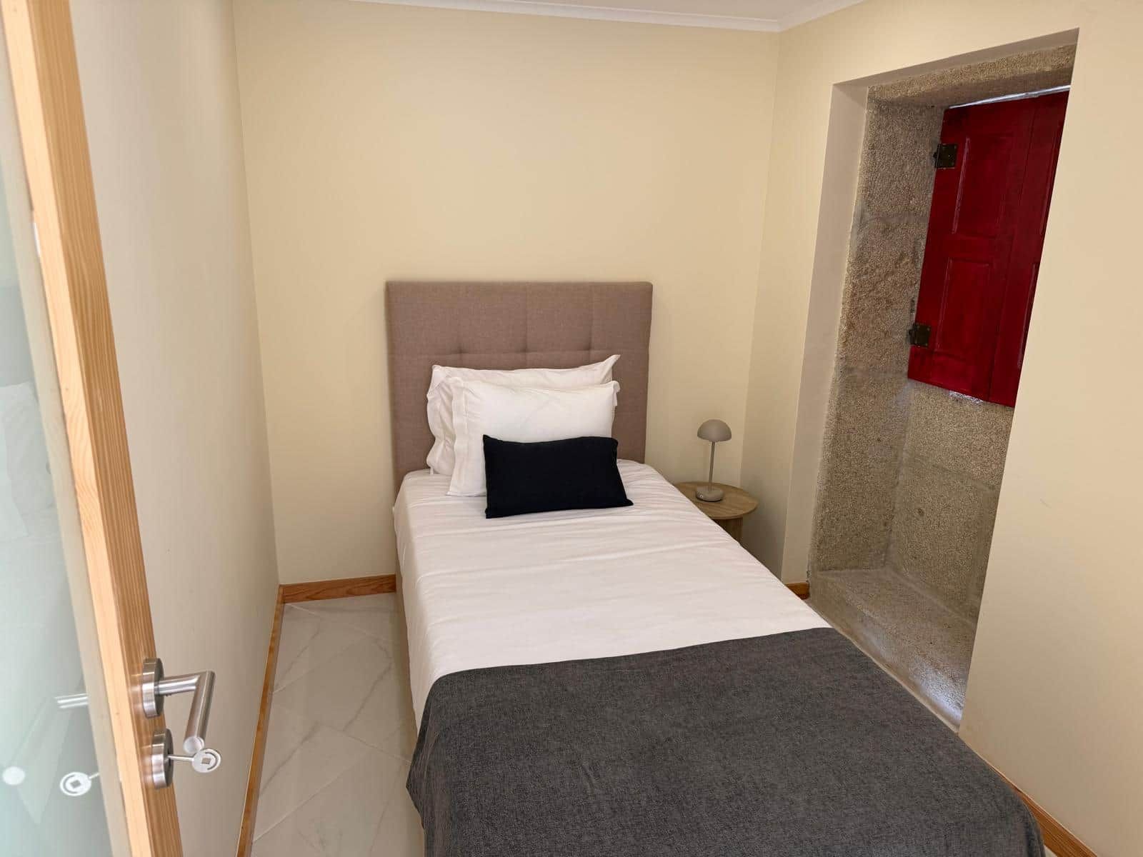 A small, neatly arranged bedroom with a bed, a bedside lamp, and a red door.