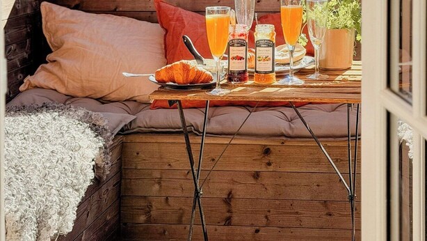 Une terrasse en bois avec une table mise pour le petit-déjeuner, comprenant du jus d’orange et de la confiture, et une vue sur les maisons au loin.