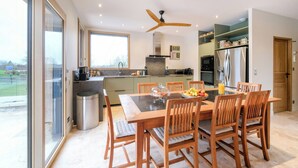 A modern kitchen with a dining table, chairs, a ceiling fan, and a view through large windows.