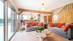 A spacious living area with two grey sofas, wooden beams, a large window, and a kitchen in the background.
