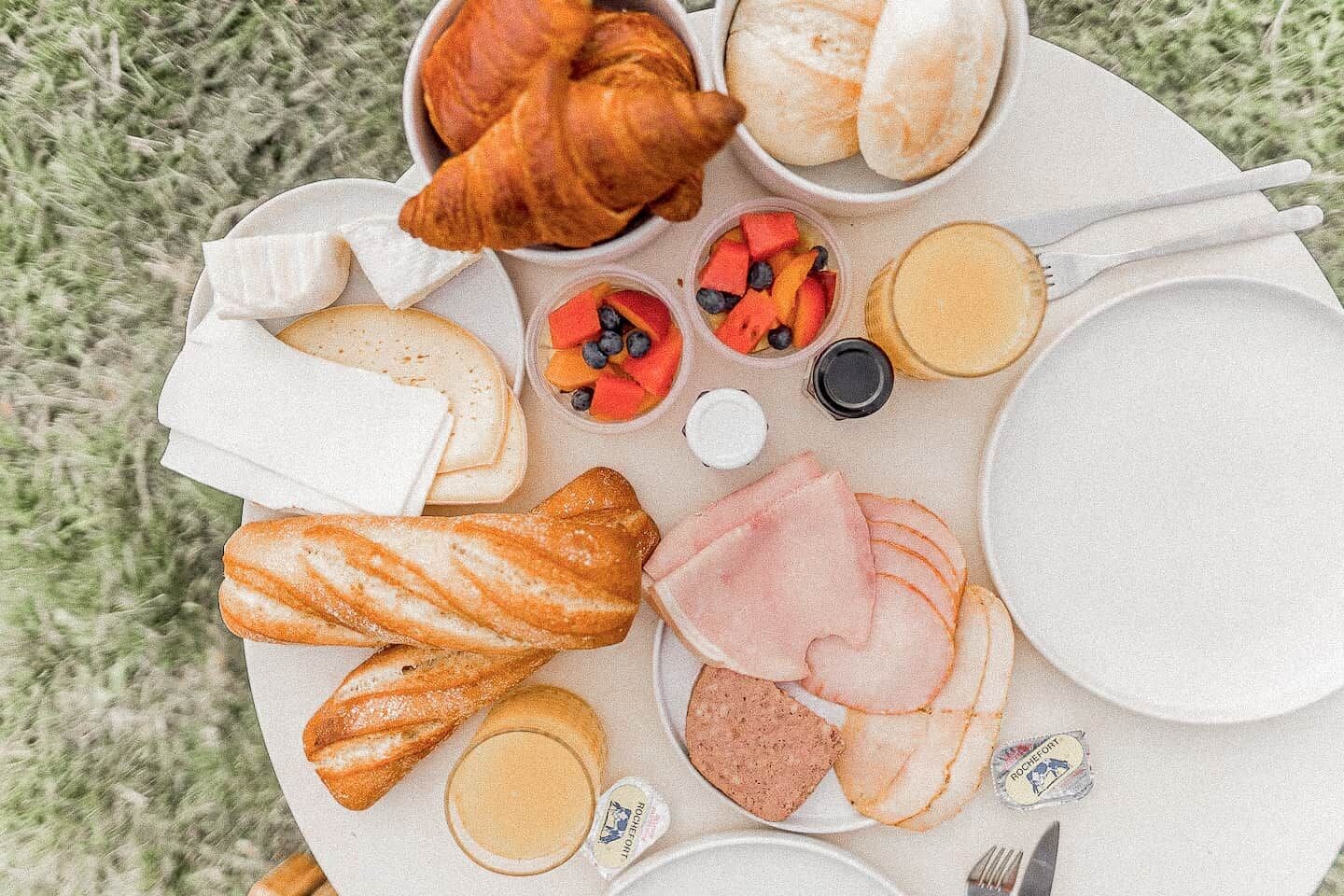 A picnic setup with bread, croissants, ham, cheese, and fruit on a white plate.