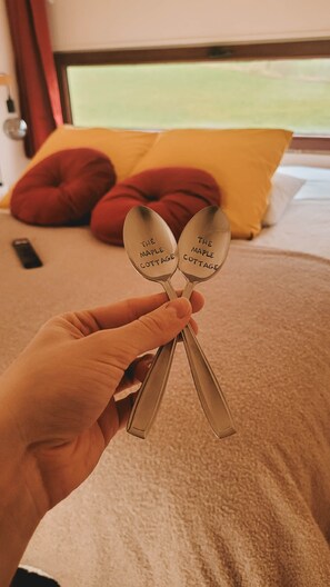 Une main tenant deux cuillères sur lesquelles est gravé le nom « The Maple Cottage », un lit avec un oreiller jaune et, à l’arrière-plan, un oreiller rouge.