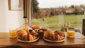 A breakfast setting with croissants, orange juice, and a bowl of fruit on a wooden table by a window. - Maple Cottage - Tiny House with private spa (Profondeville)