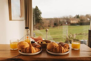 Aamiaispöytä, jossa on croissantteja, appelsiinimehua ja kulho täynnä hedelmiä, ja pöytä on sijoitettu ikkunan viereen olevalle puupöydälle.
