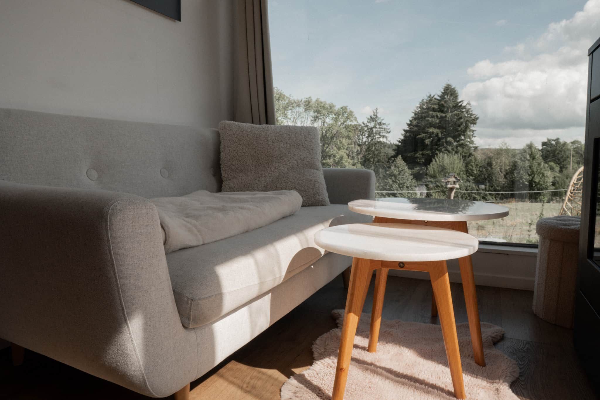 A modern living room with a grey sofa, two white side tables, and a view of trees through a window.