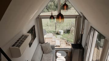 Habitación espaciosa con techo alto, unidad de aire acondicionado integrada, sofá, una mesita y vista a un jardín a través de un ventanal grande.