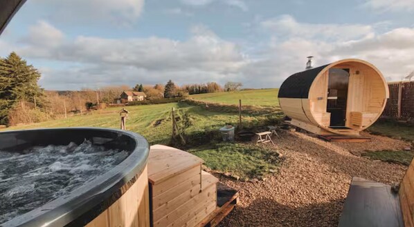 Un bain à remous, un sauna en bois et un chalet en bois avec une porte ouverte, le tout situé dans un paysage rural sous un ciel dégagé.