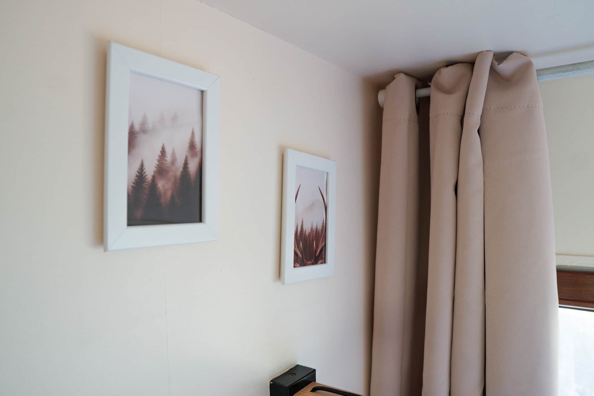 Una habitación con dos cuadros en la pared, una ventana con cortinas color beige y un objeto negro pequeño sobre una superficie de madera.