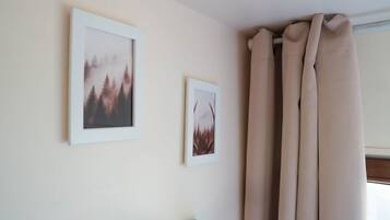 Una habitación con dos cuadros en la pared, una ventana con cortinas color beige y un objeto negro pequeño sobre una superficie de madera.
