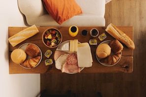 Una mesa de madera con pan, queso, jamón, croissants y un bol de fruta.